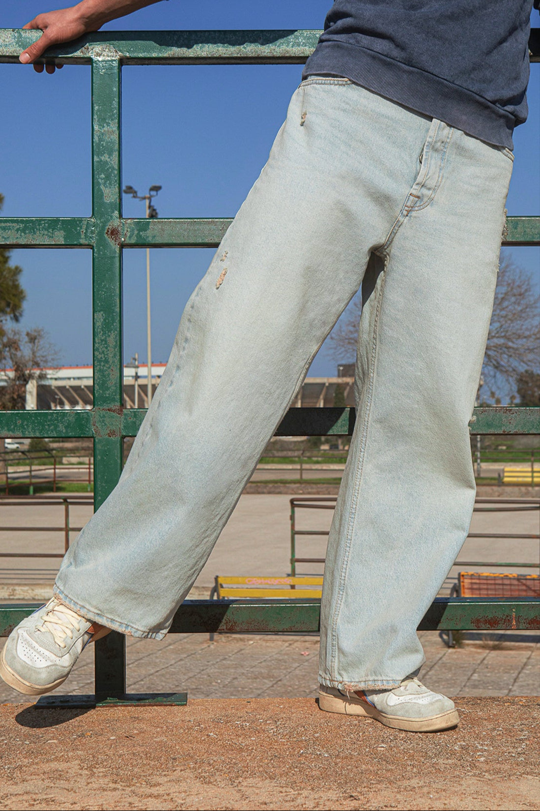 Person wearing light blue jeans and white sneakers standing on a concrete surface with a green metal railing and blue sky in the background.slider_item_dpAJnJ