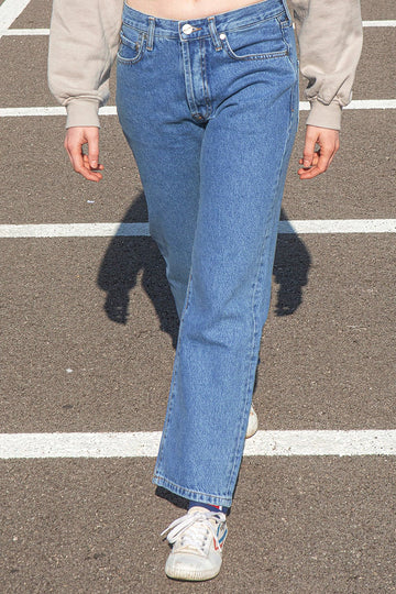 Person wearing a beige sweatshirt and KLSH RYAN faded blue jeans walking on a road.