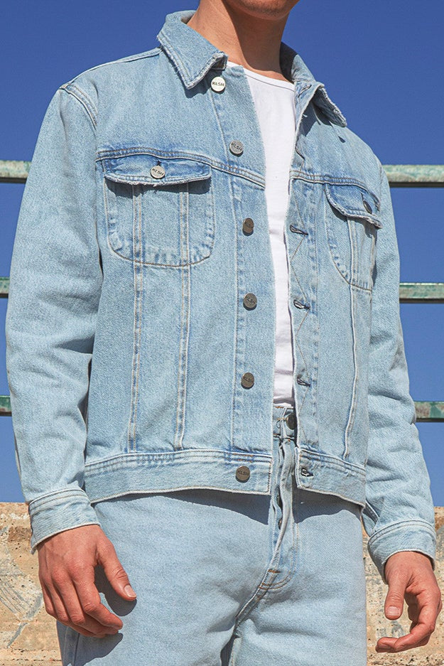 Person wearing a light blue denim jacket and jeans standing against a graffiti-covered wall.