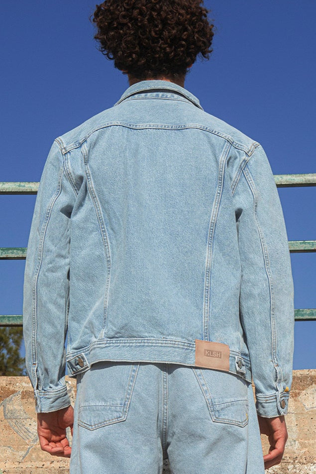 Person wearing a light blue denim jacket with a visible brand label against a clear blue sky.