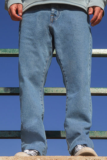 Person wearing faded blue jeans and a gray hoodie standing on a metal railing against a clear blue sky.