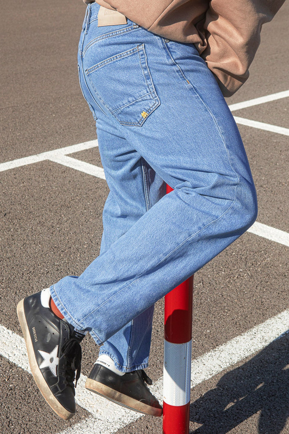 Person wearing blue jeans and black shoes standing on a red and white striped pole in an outdoor setting.