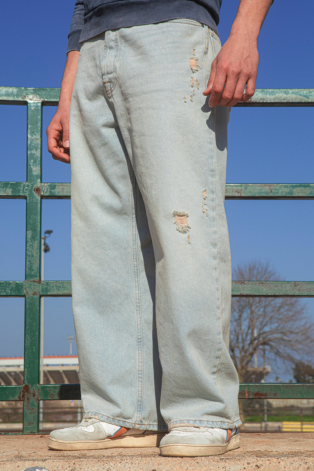 Person wearing light blue jeans with slight distressing, standing against a green metal fence with a clear blue sky.