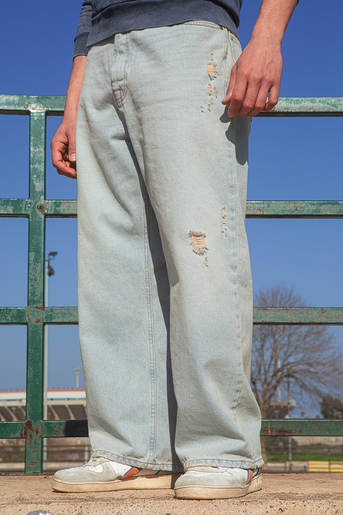 Person wearing light blue jeans with slight distressing, standing against a green metal fence with a clear blue sky.