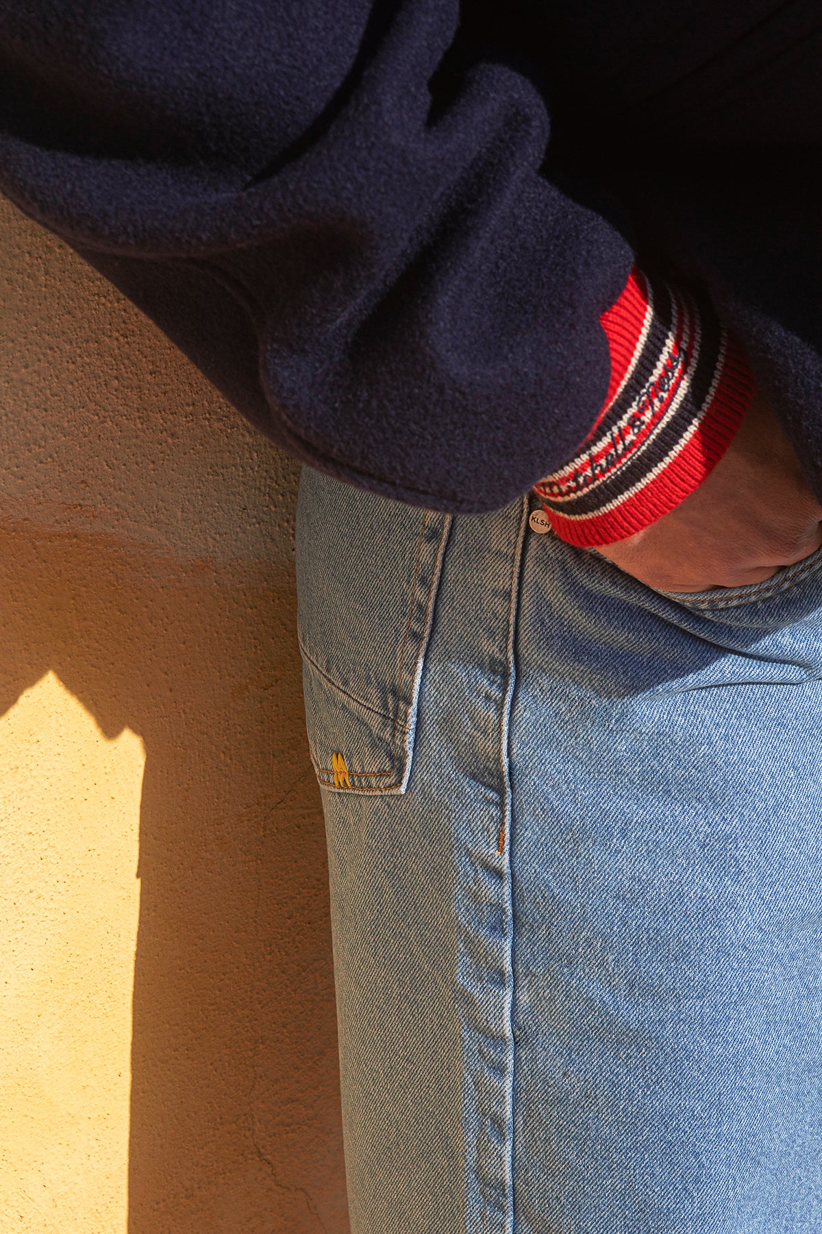 Close-up of a person wearing a navy sweater with a red and white patterned cuff, blue jeans, and a beige wall in the background.