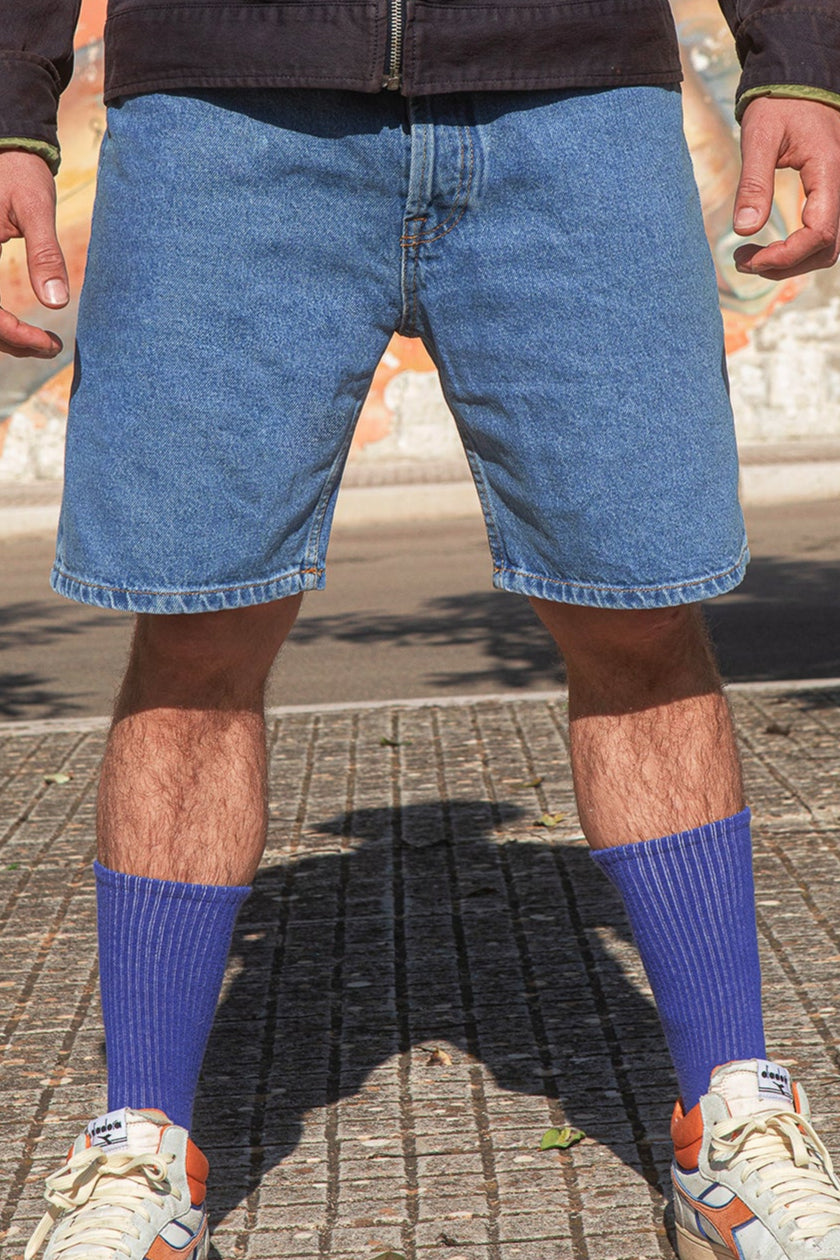 Person wearing blue denim shorts, purple socks, and white sneakers on a street.
