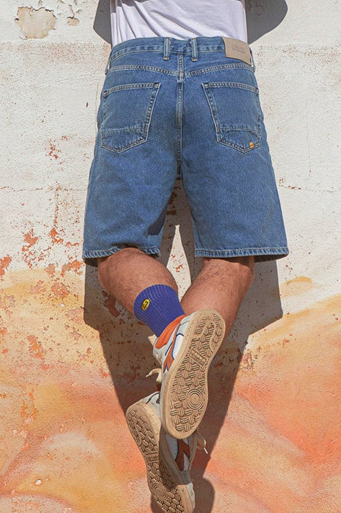 Person wearing blue denim shorts, purple socks, and white sneakers on a street.