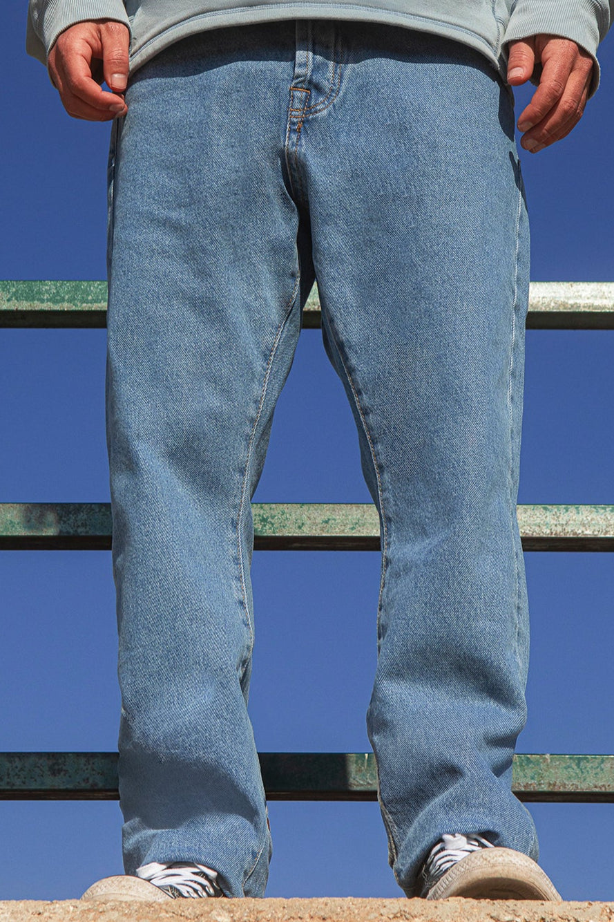 Person wearing faded blue jeans and a gray hoodie standing on a metal railing against a clear blue sky.