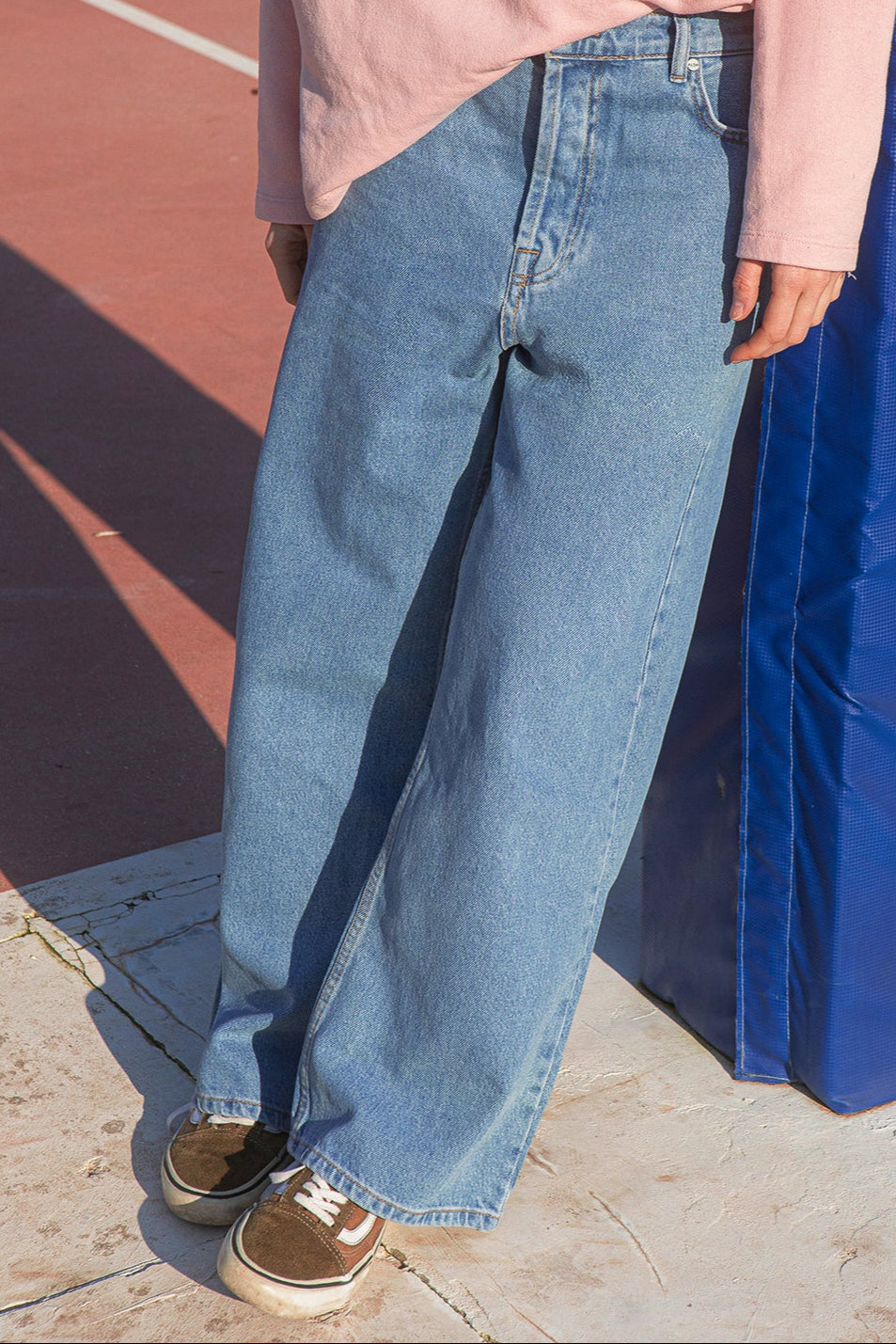 Person wearing a light pink shirt and blue jeans standing on a concrete surface with a blue wall in the background.
